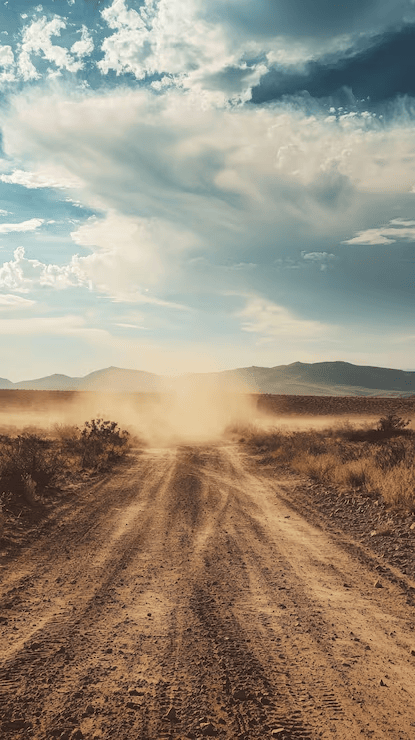 A dusty dirt road stretches into the distance, surrounded by sparse vegetation and distant mountains under a partly cloudy sky.