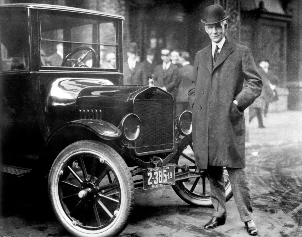 A man in a coat and bowler hat stands beside an early 20th-century car, with a crowd in the background.