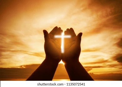 Silhouette of hands holding a glowing cross against a sunset sky.